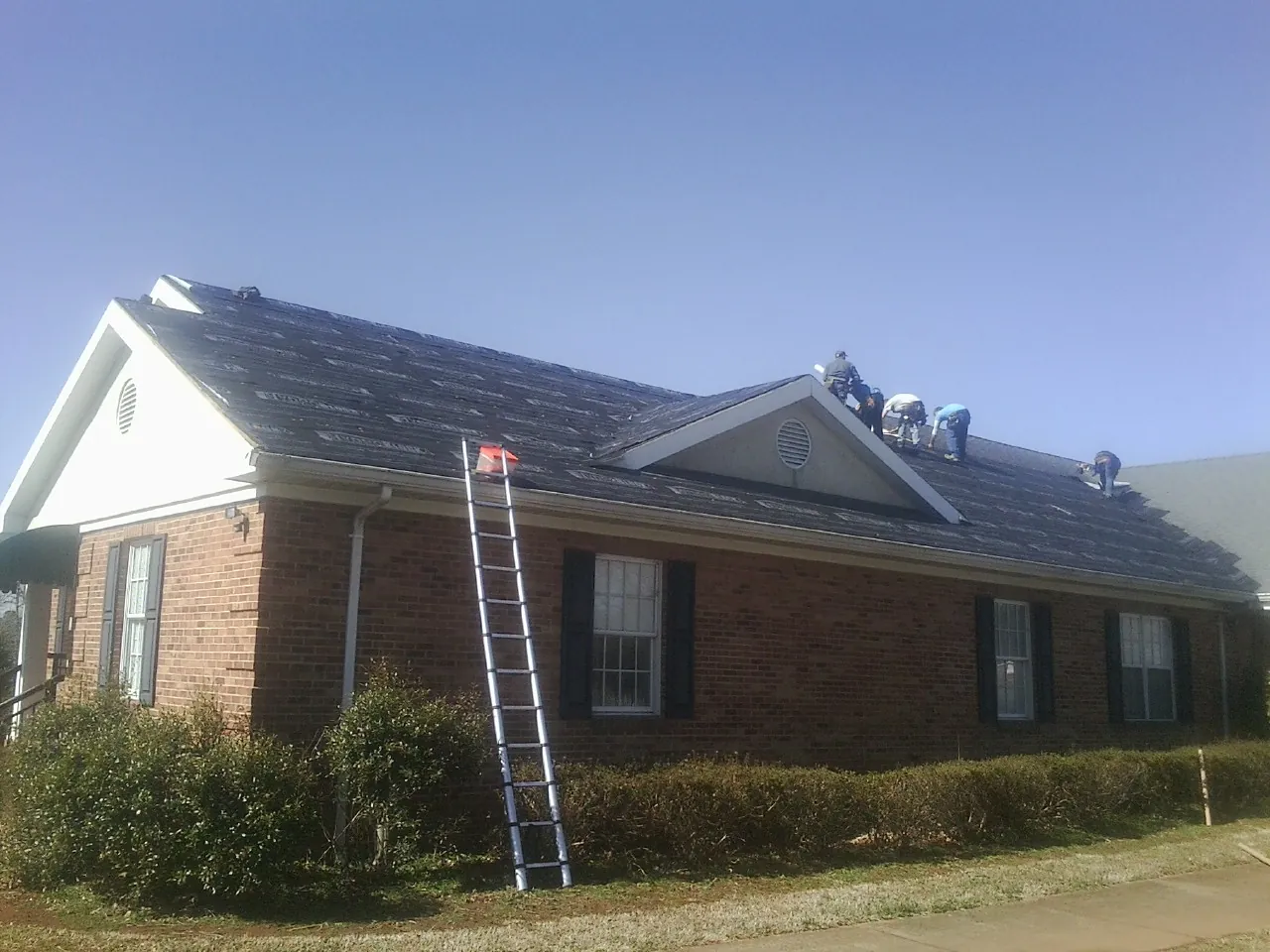 Crew tearing shingles off a brick ranch roof with a ladder set — Metal Roof Repair work by our crew in Kirkville