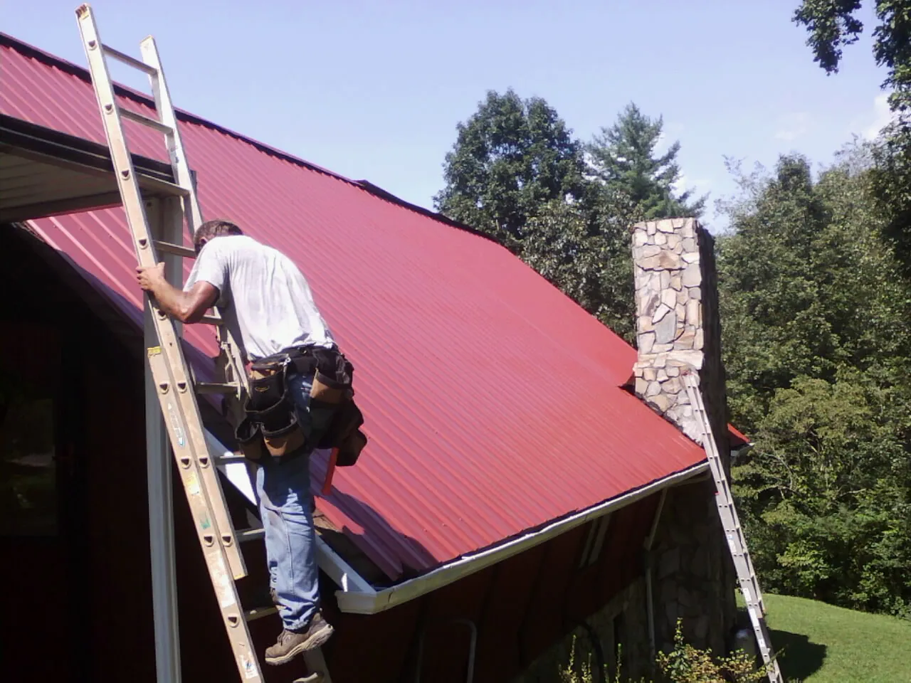Roofer on a ladder beside a red metal roof and stone chimney — Soffit & Fascia Repair by Roofing Brothers in Kirkville